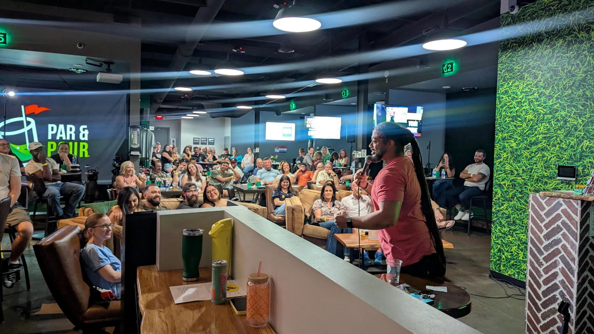 A packed comedy night in a bar: a comedian on the microphone and a full room of people watching from couches, bar stools and high-tops.