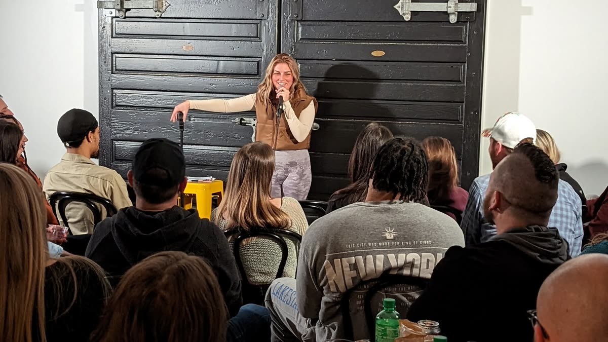 A comedian on the microphone in a small room, the audience seated close and filling every chair.