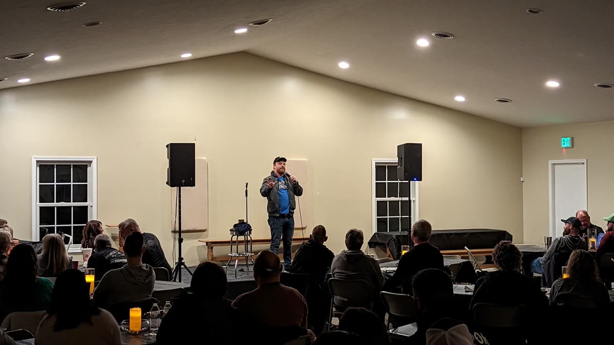 A comedian on a low stage in an event hall, playing to a seated crowd at candlelit tables.