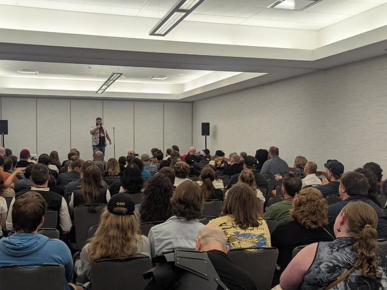 A comedian performing to a large seated audience filling every chair in the room.