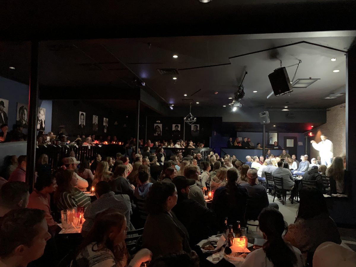 A packed, candlelit room of tables watching a comedian under a spotlight.