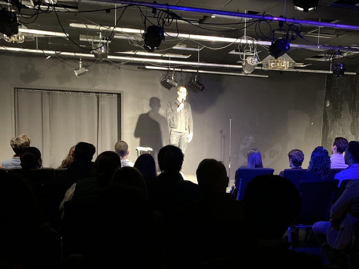 A comedian under stage lights in a black-box room, the audience seated in rows.