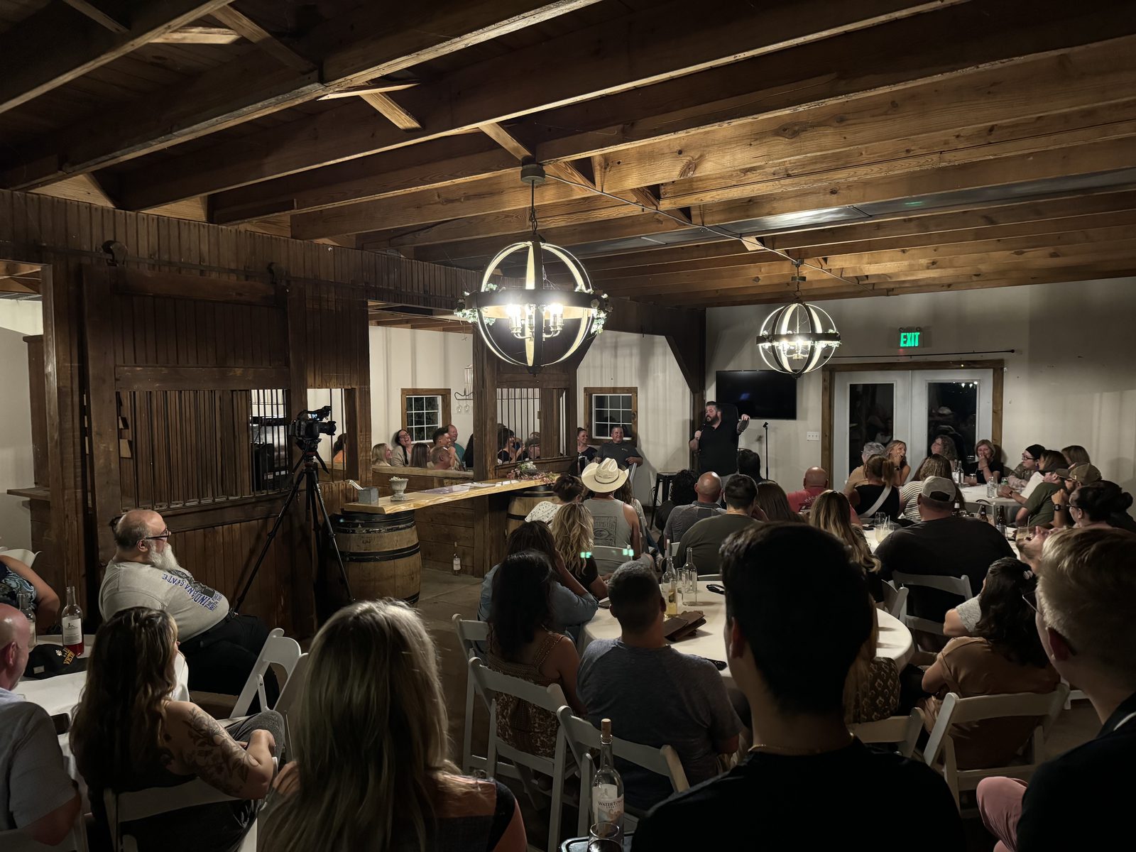 A packed barn venue with wooden beams and chandeliers, the crowd at tables watching a comedian.